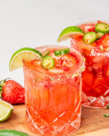 Two strawberry jalapeno margaritas topped with jalapeno slices and lime wedges.