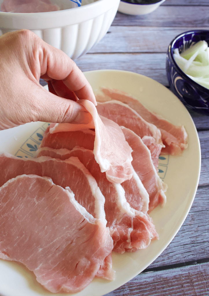 A hand holding up a thin slice of raw pork.