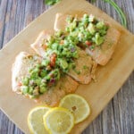 Baked salmon on a wooden cutting board topped with avocado pico de gallo.