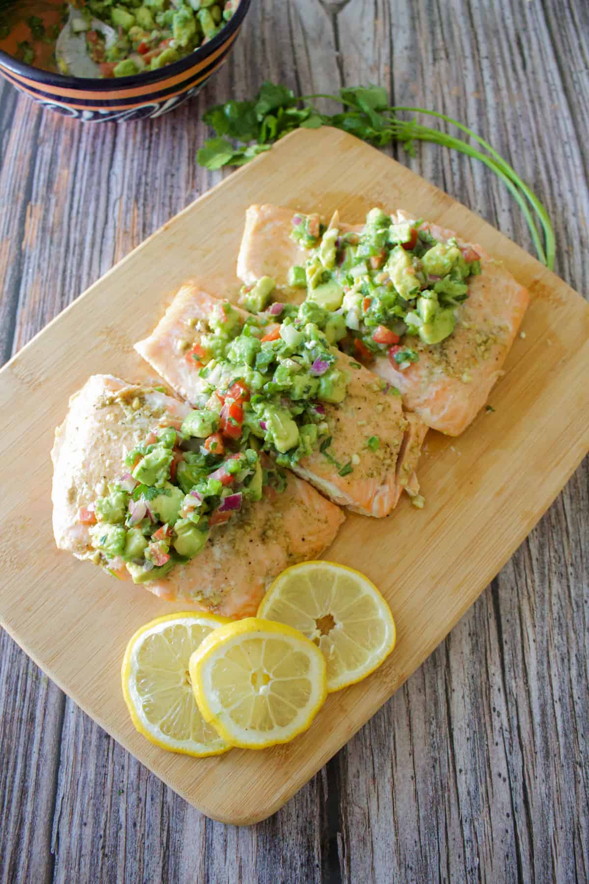 Baked salmon on a wooden cutting board topped with avocado pico de gallo.