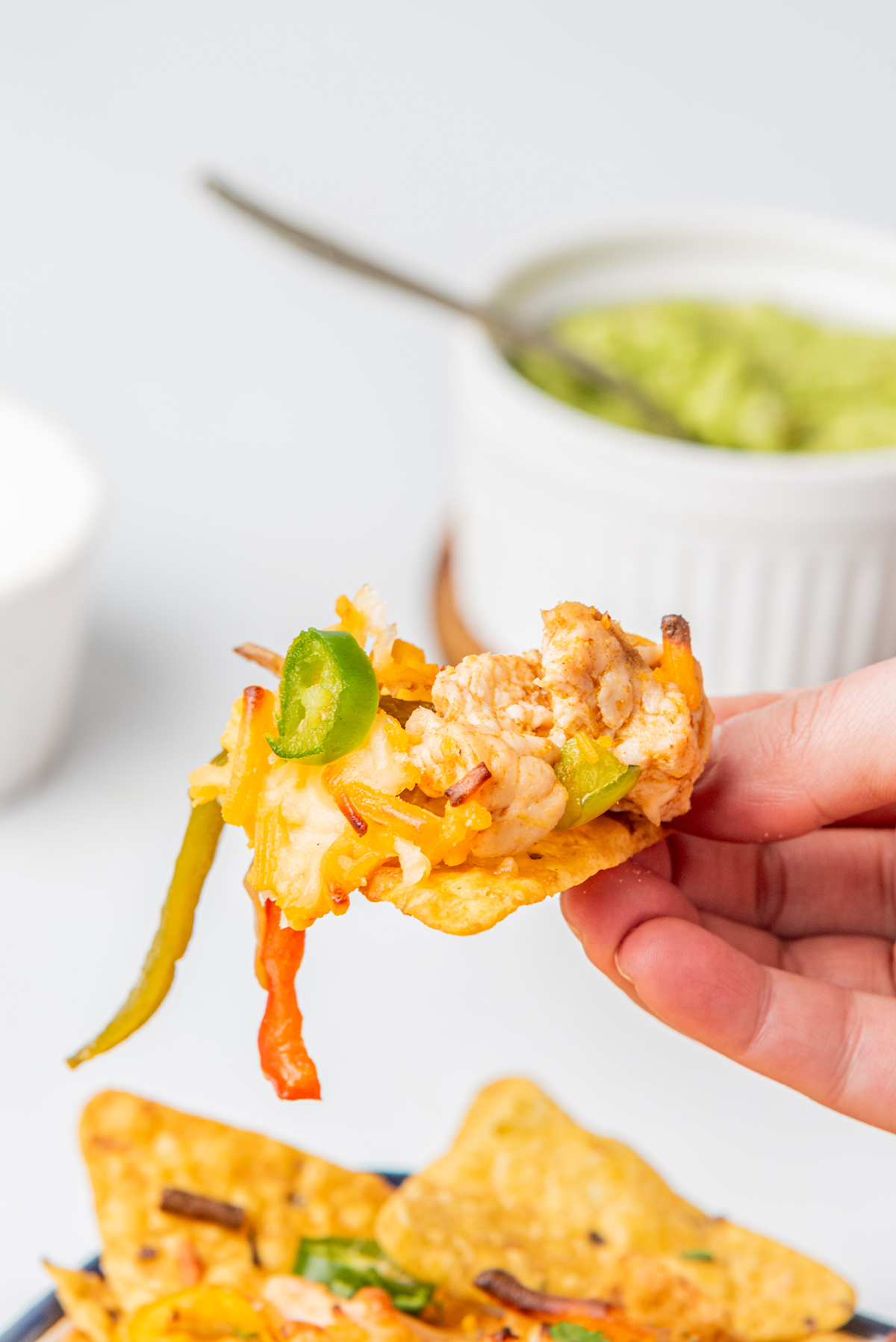 A hand holding a tortilla chip with chicken nacho toppings.