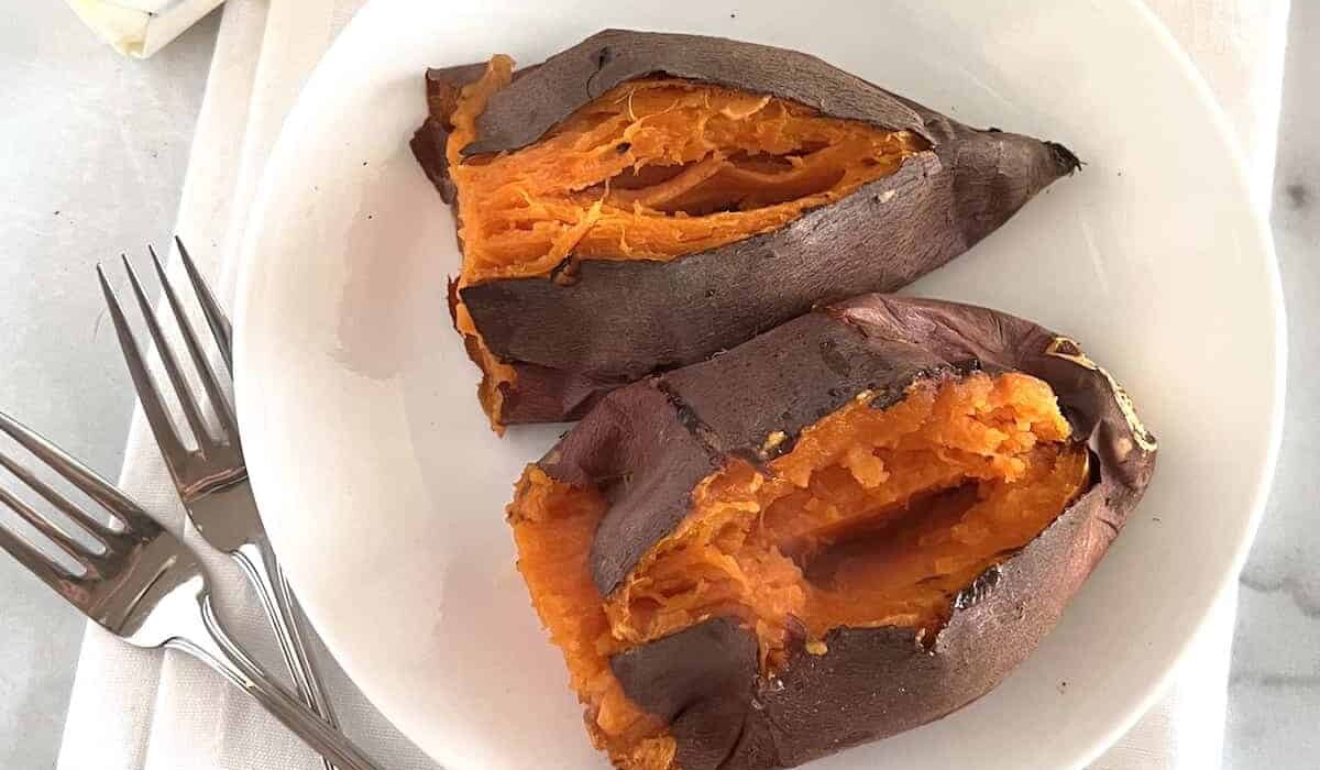 A white plate with two baked sweet potatoes split open, revealing their orange, soft interiors. Two forks are placed beside the plate on a white napkin.