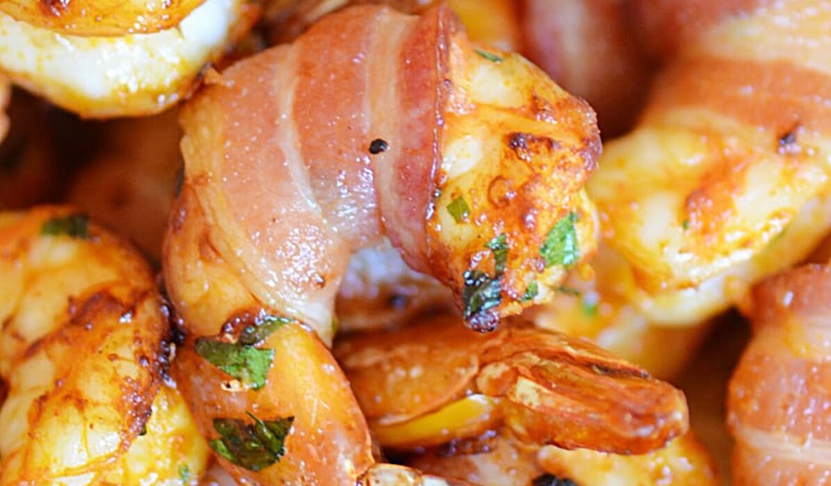 Close-up of shrimp wrapped in crispy bacon, garnished with herbs. The shrimp appear golden and juicy, suggesting they are freshly cooked and well-seasoned.