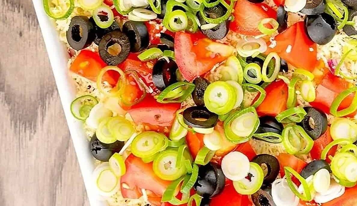 A close-up of a layered salad featuring chopped tomatoes, sliced black olives, and chopped green onions on top of a rectangular dish. The vibrant colors contrast against a wooden table background.