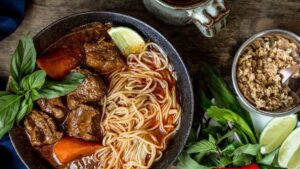 A bowl of noodle soup with beef chunks, carrots, and a lime wedge, garnished with fresh basil. Next to it, a small dish holds crushed peanuts, and another plate features lime wedges, basil leaves, and red chilies.