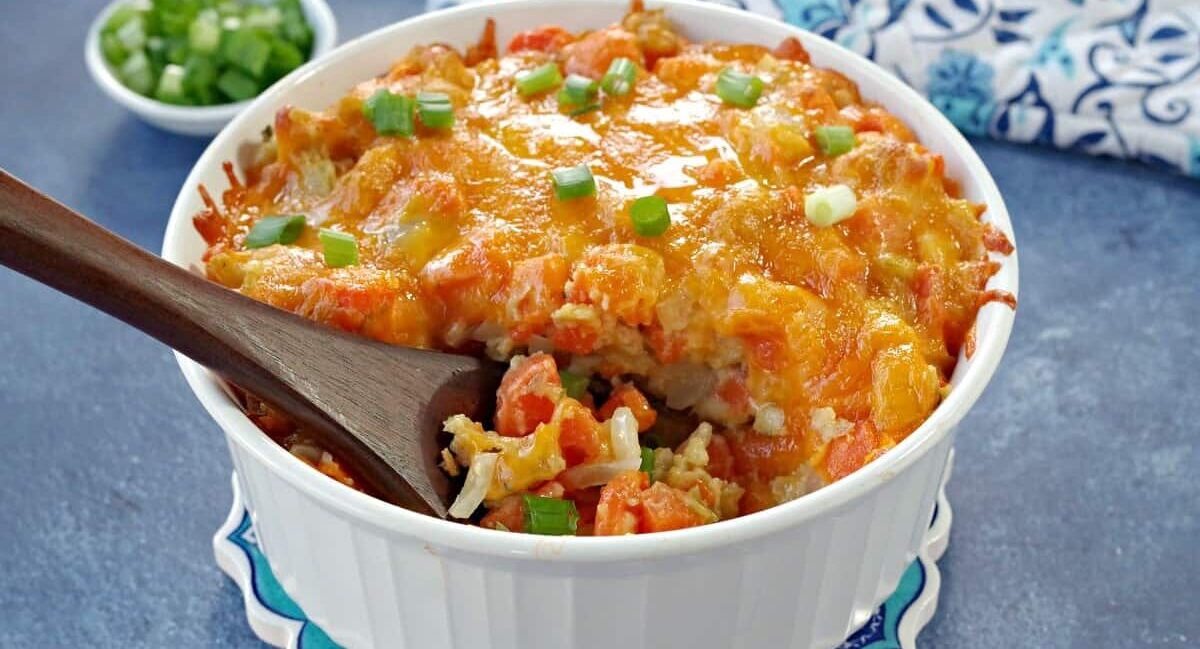 A baked casserole in a white dish topped with melted cheese and chopped green onions. A wooden spoon rests in the dish. A small bowl with chopped green onions is in the background on a blue surface.