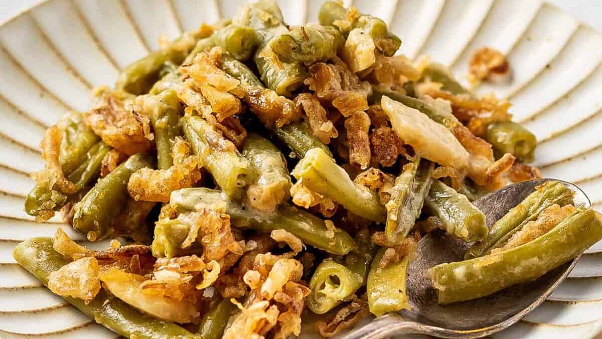 A plate of green bean casserole topped with crispy fried onions. The green beans are mixed with a creamy sauce and are richly textured. A spoon rests on the plate, partially dipped into the dish.