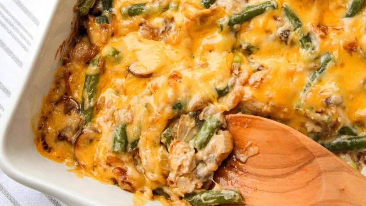 A dish of green bean casserole with melted cheese and caramelized onions, partially served with a wooden spoon, in a white baking dish on a cloth background.