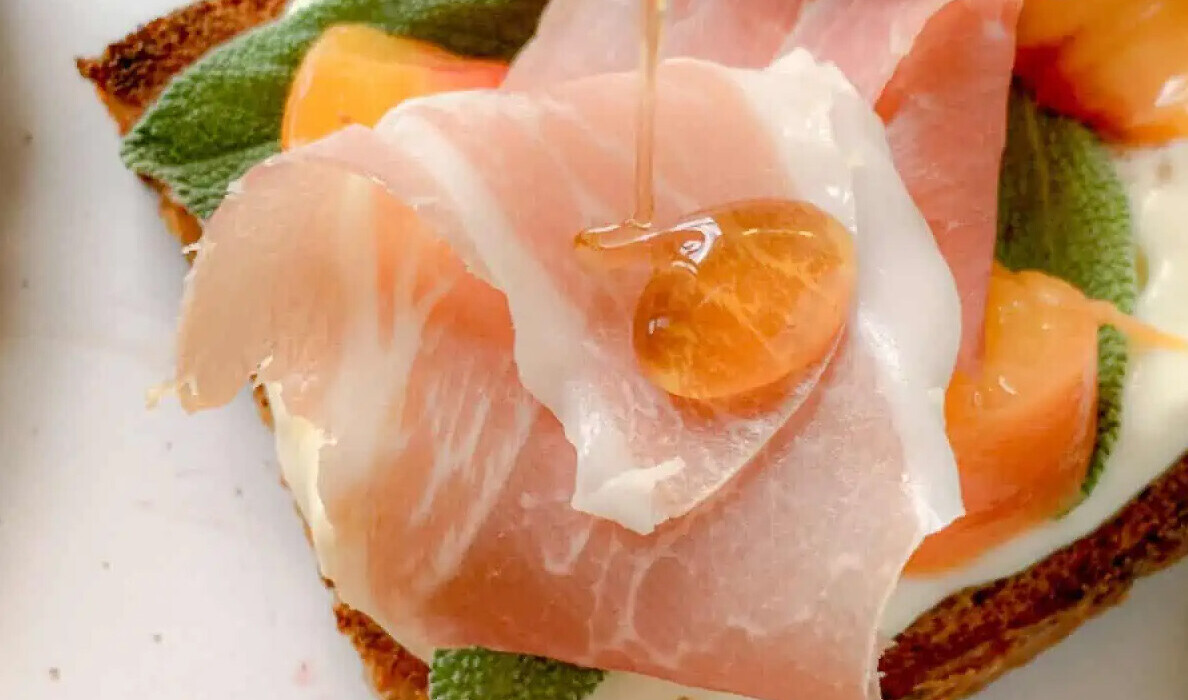 Close-up of a slice of toast topped with prosciutto, sage leaves, and peach slices. Honey is being drizzled over the prosciutto, creating a glossy droplet. The combination of ingredients creates an appetizing presentation.