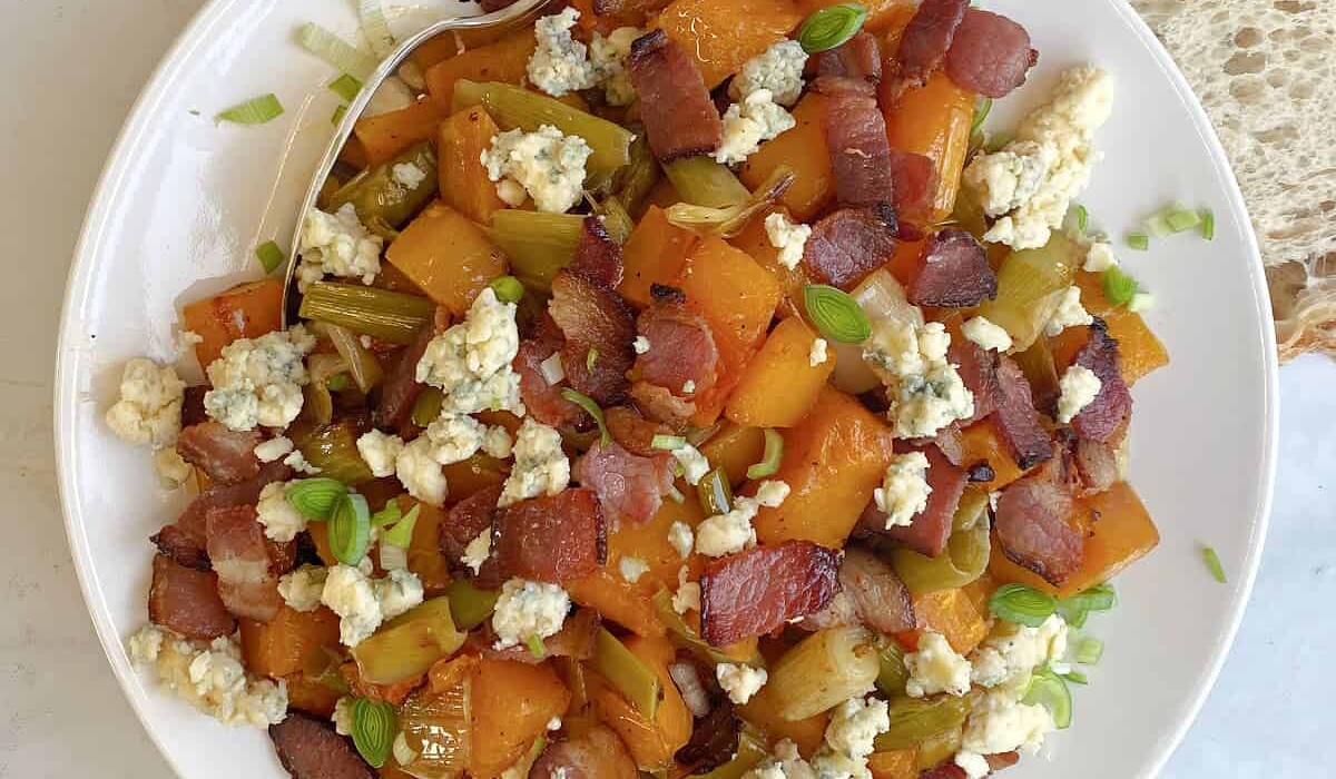 A plate of roasted butternut squash and celery, topped with crispy bacon, crumbled blue cheese, and garnished with fresh green onions. A spoon rests on the side, and there's a slice of bread nearby.