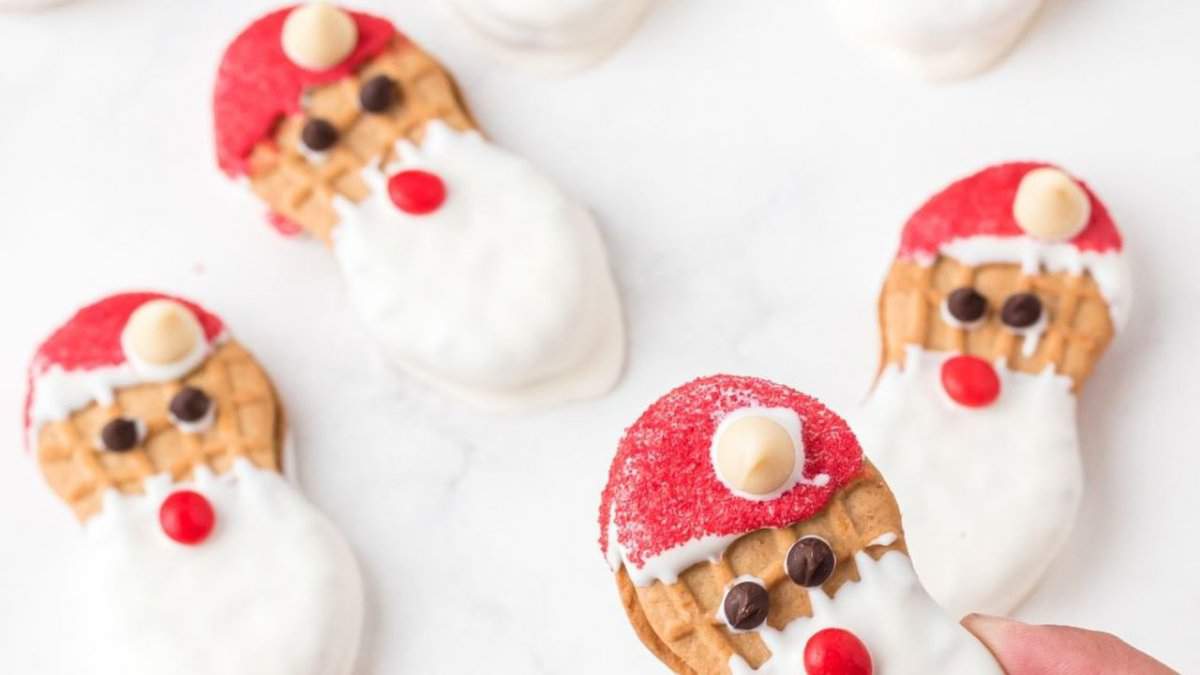 Peanut-shaped cookies decorated as Santa faces with red sugar hats, white chocolate beards, mini chocolate chips for eyes, red candy noses, and white chocolate chips for hat tops, arranged on a white surface.