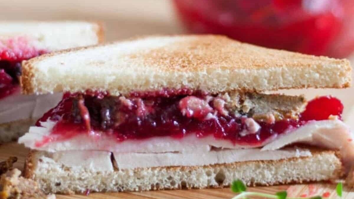 A close-up of a sliced turkey sandwich with cranberry sauce on toasted white bread. The filling is visible, showing layers of turkey and bright red cranberry sauce. The sandwich is on a wooden surface.