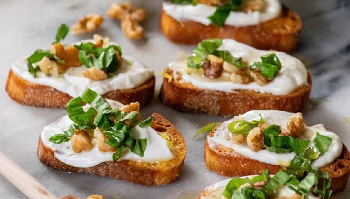 Toasted bread slices topped with creamy cheese, fresh basil, and walnuts, arranged on a marble surface.