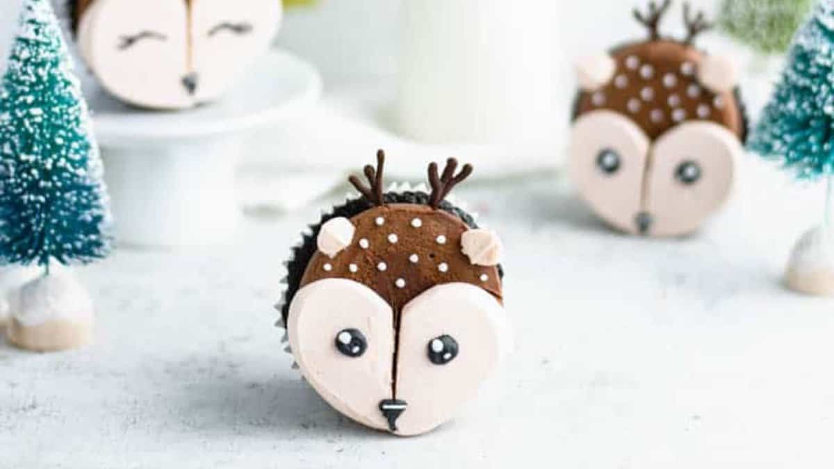 A cupcake decorated to look like a cute woodland deer with big eyes and antlers, surrounded by small, artificial snow-covered trees. Another similarly decorated cupcake is in the background, creating a whimsical forest scene.