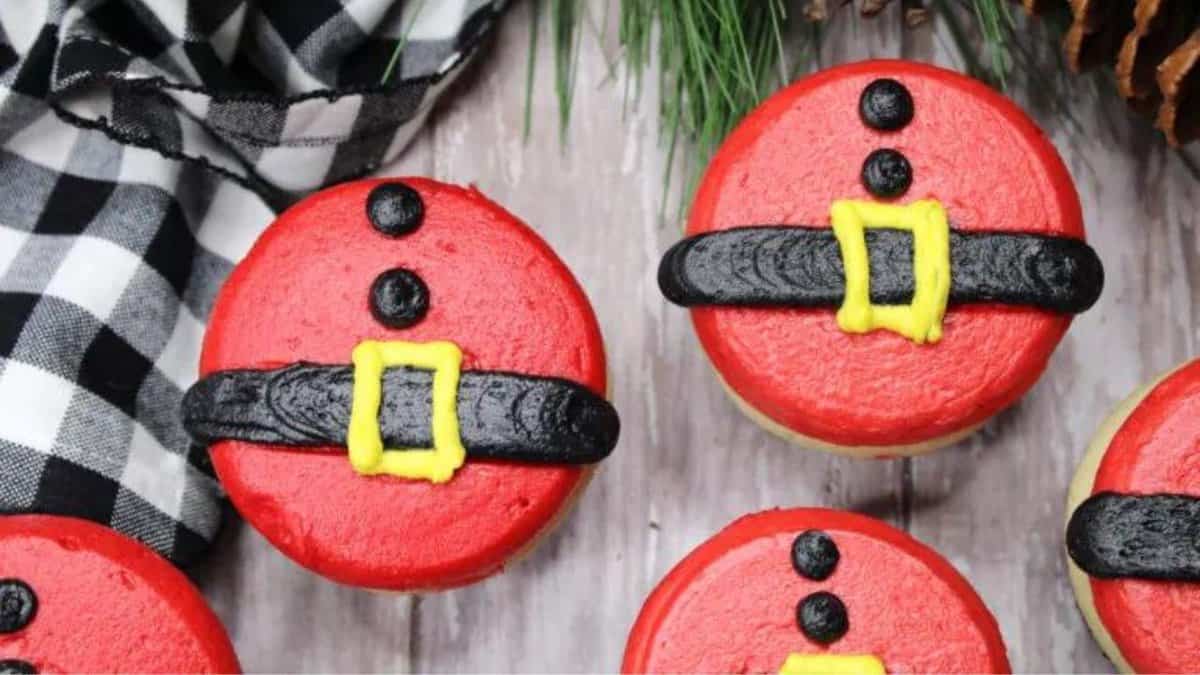 Cupcakes decorated with red icing, black “belts,” and yellow “buckles” to resemble Santa’s suit, arranged on a wooden surface next to pine branches and a black-and-white checkered cloth.