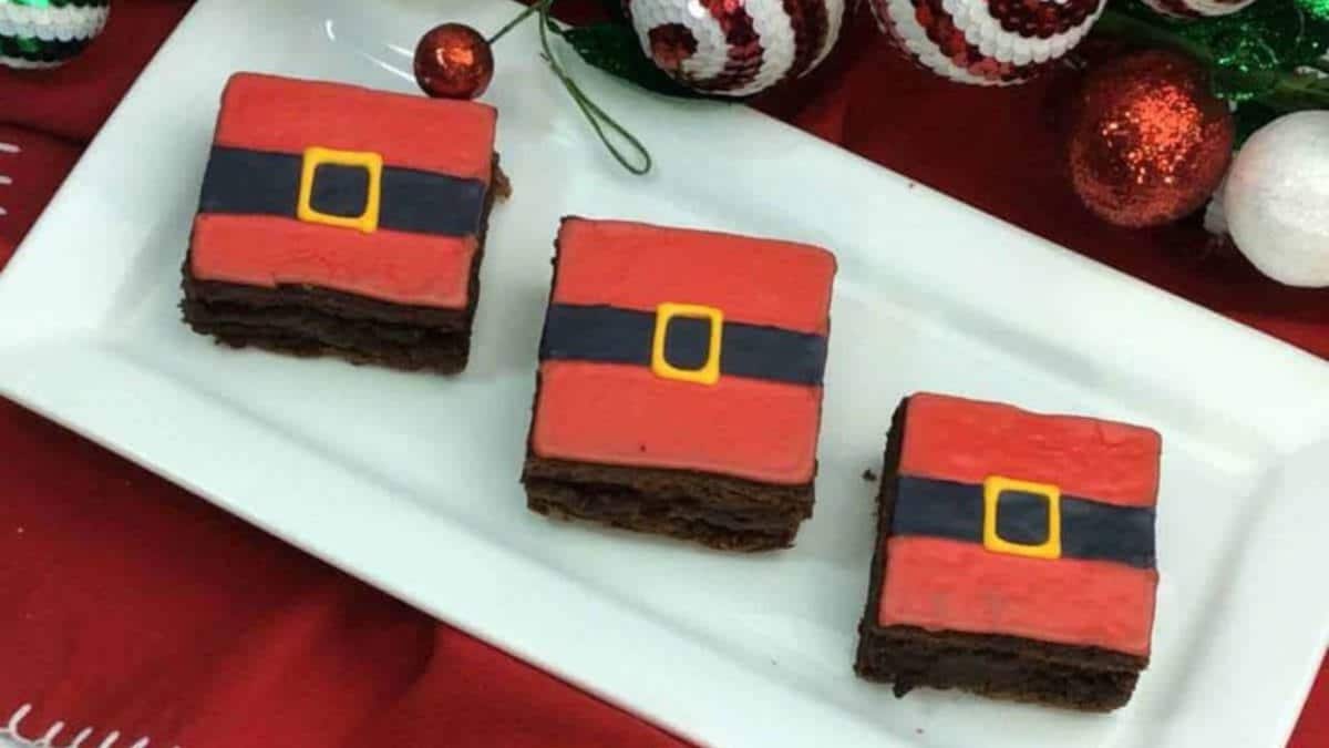 Three brownies decorated to look like Santa’s belt, with red icing, a black stripe, and a yellow buckle, are arranged on a white rectangular plate. Festive holiday decorations are in the background.