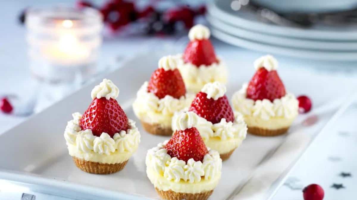 Six mini cheesecakes topped with strawberries decorated as Santa hats using whipped cream are arranged on a white plate with a candle and plates in the blurred background.