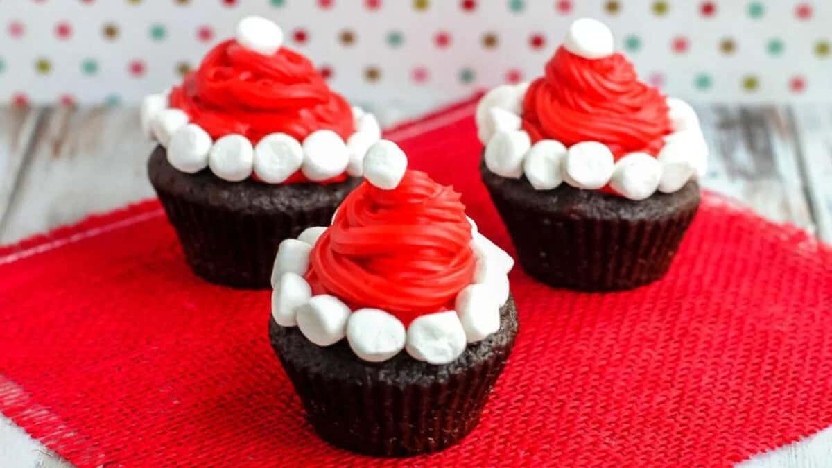 Three chocolate cupcakes topped with red frosting shaped like Santa hats, each decorated with mini marshmallows as hat trim and a white marshmallow on top, displayed on a red cloth with a festive background.