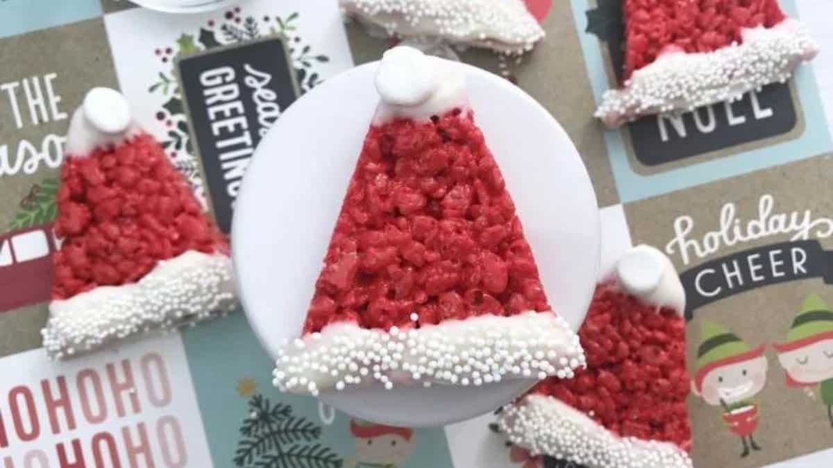 Five triangle-shaped Rice Krispies treats decorated as Santa hats with red coloring, white frosting on the base, a frosting pom-pom at the tip, and white sprinkles, arranged on festive holiday-themed paper.