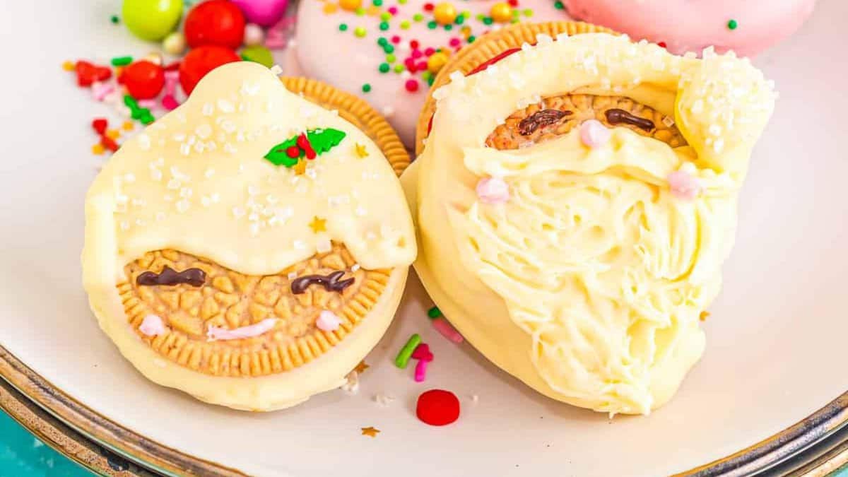 Two vanilla sandwich cookies decorated with white chocolate and candy to resemble cute holiday faces, one with a holly leaf and the other with a beard, displayed on a plate with colorful sprinkles and candies nearby.