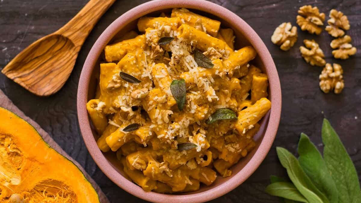 A bowl of pasta topped with a creamy pumpkin sauce, garnished with grated cheese and sage leaves. It's surrounded by walnuts, a wooden spoon, fresh sage leaves, and a slice of pumpkin on a dark wooden surface.