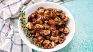 A white bowl filled with cooked, sliced mushrooms garnished with a sprig of fresh thyme, placed on a turquoise surface next to a white and blue checkered cloth.