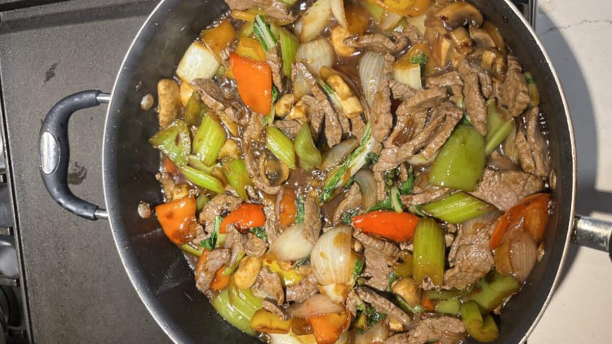 A stir-fry dish in a pan featuring sliced beef, bell peppers, onions, mushrooms, and green leafy vegetables. The ingredients are coated in a dark sauce, with a mix of green, red, and brown tones.