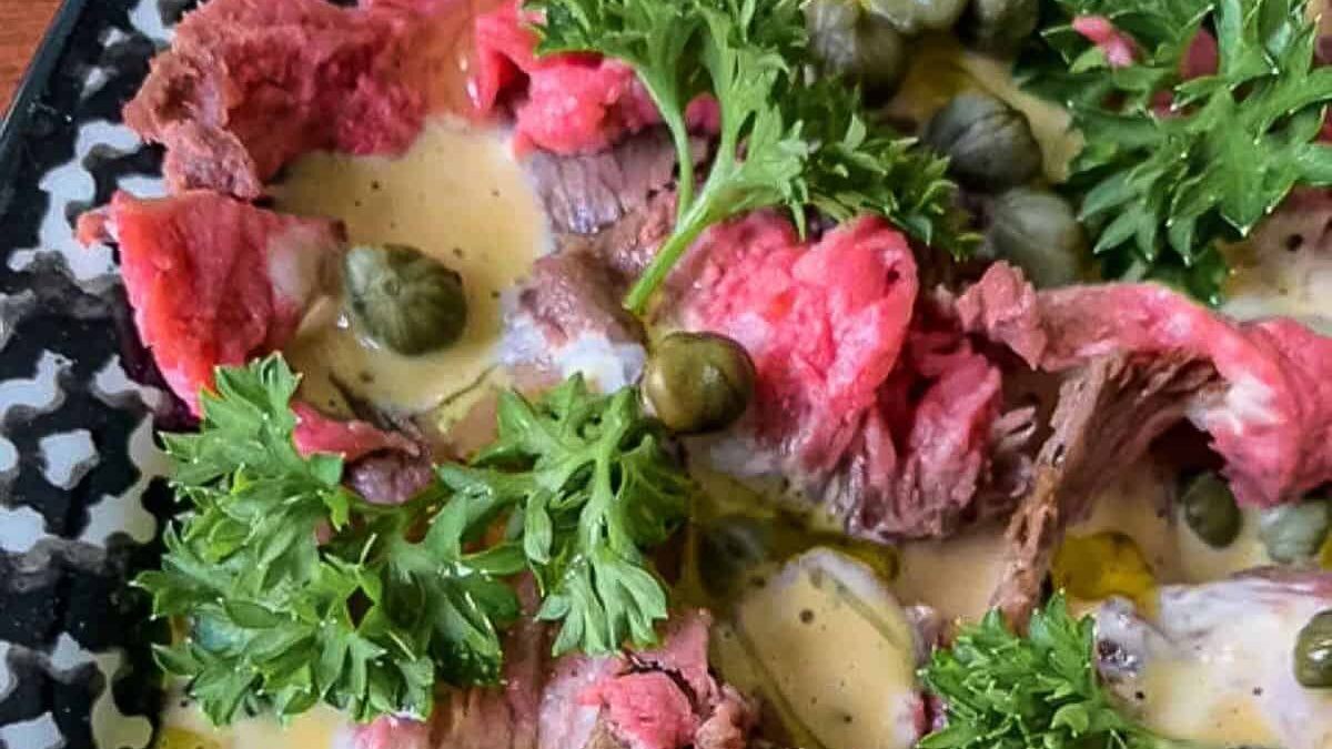 Close-up of a dish featuring thin slices of rare beef topped with capers and fresh parsley, drizzled with a creamy sauce, all arranged on a patterned black and white plate.