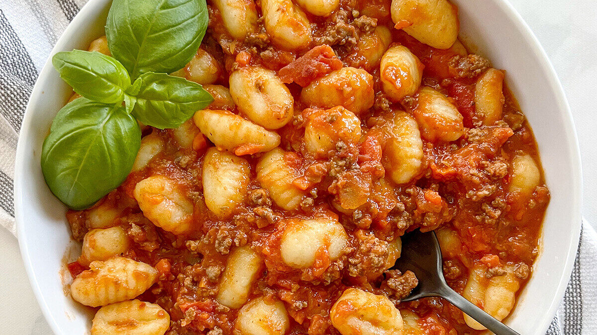 A bowl of gnocchi is topped with a rich tomato and meat sauce, accompanied by fresh basil leaves. A fork rests in the dish, displaying the hearty, savory meal.