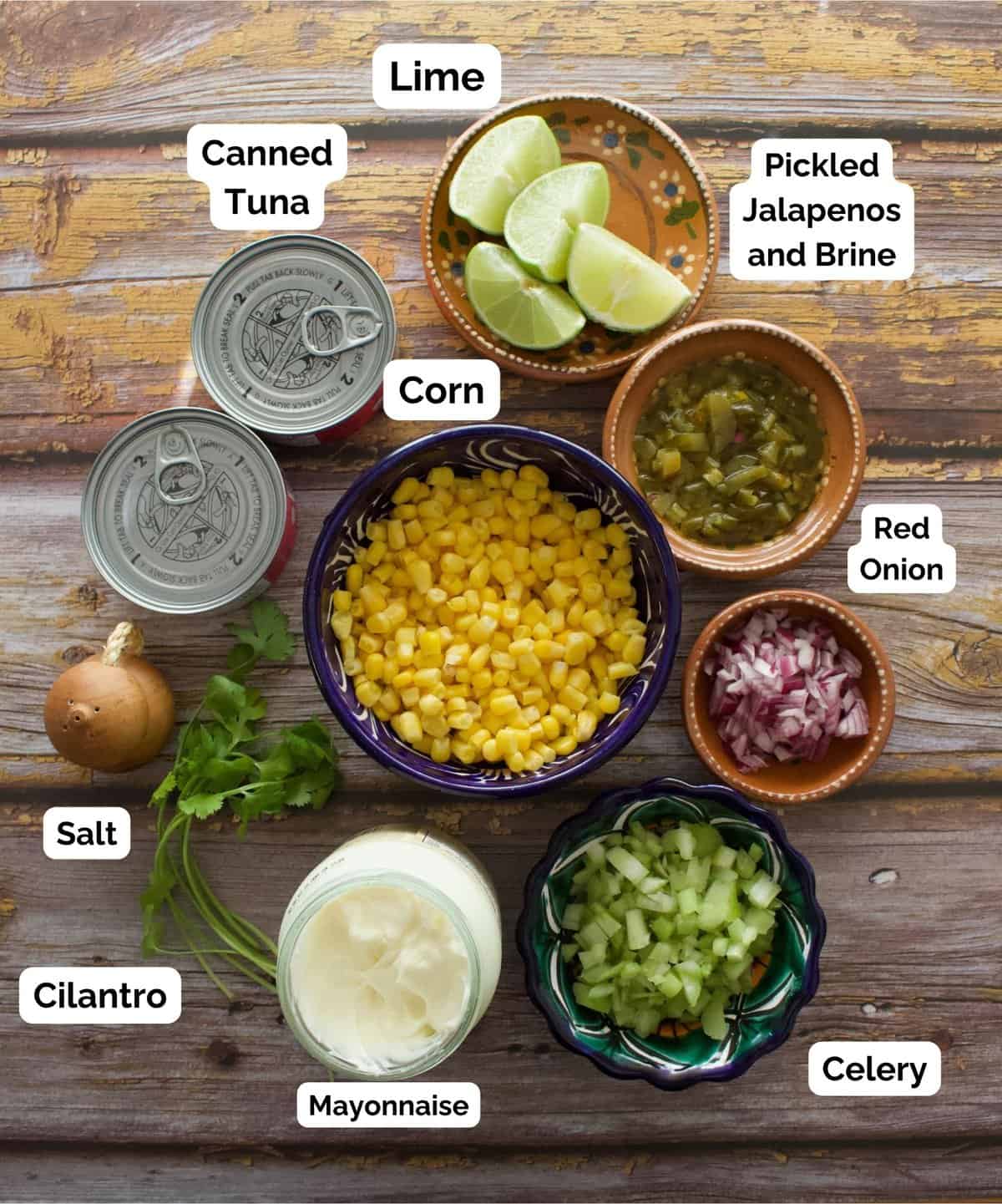 The ingredients to make the Corn Tuna Salad labeled and sitting on a wooden surface.
