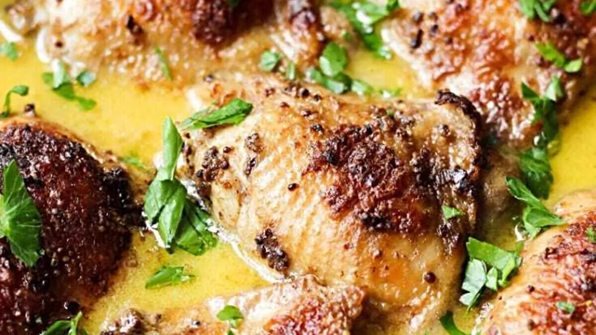 Close-up of crispy chicken pieces simmering in a creamy sauce, garnished with fresh parsley. The dish features golden-brown skin and specks of ground black pepper, all nestled in a rich, flavorful broth.