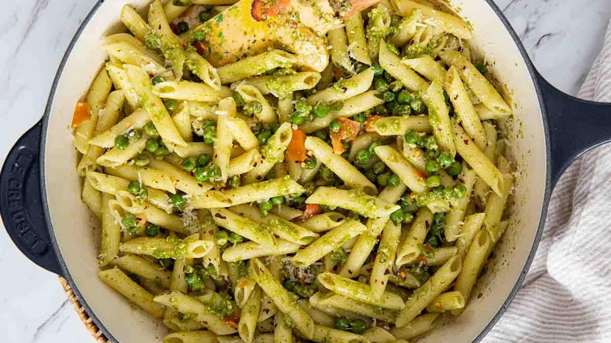 A pot filled with penne pasta mixed with green peas, chopped carrots, and stirred with a creamy pesto sauce. A wooden spatula rests in the pasta, and the pot is placed on a marbled countertop with a cloth nearby.