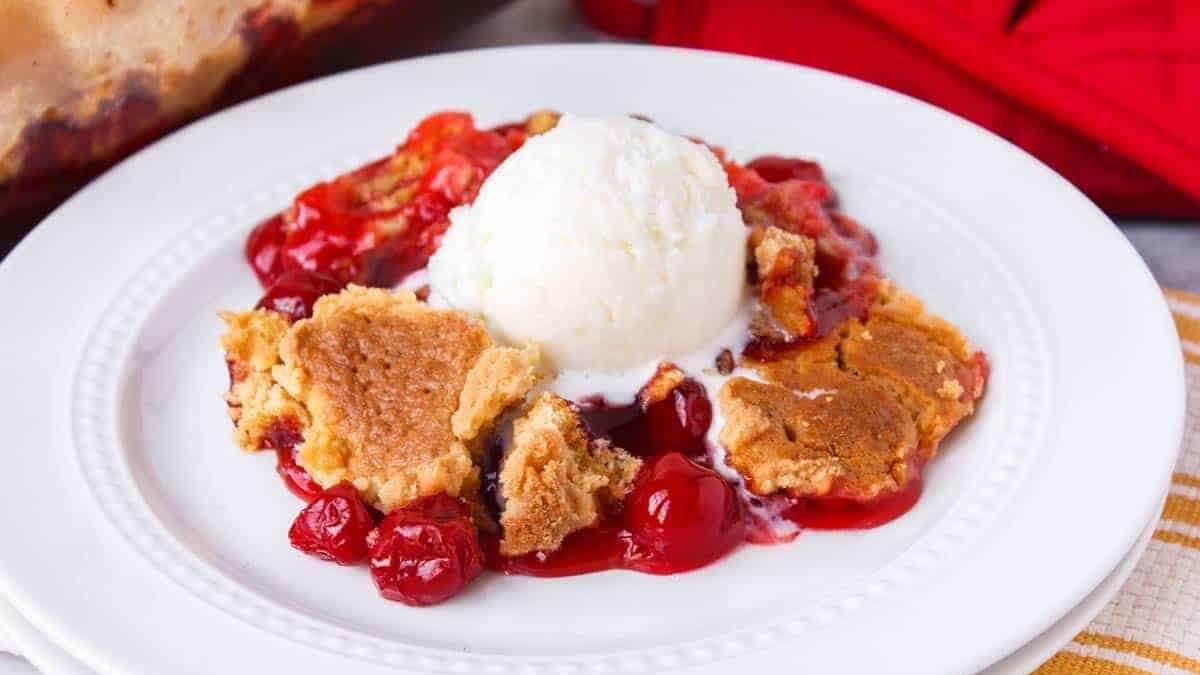 A plate of cherry cobbler topped with a scoop of vanilla ice cream. The cobbler features a golden-brown crust with visible cherry filling. A portion of a baking dish and a red towel are in the background.