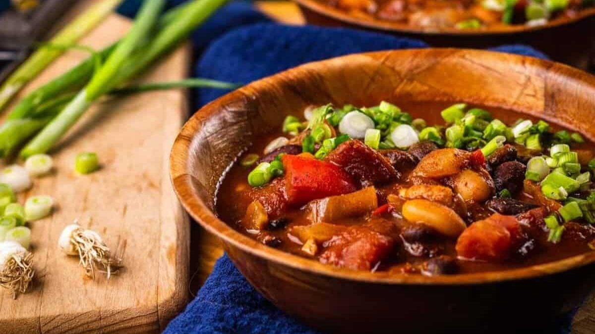 A wooden bowl filled with hearty chili, featuring beans, tomatoes, and green onions for garnish. Fresh green onions are on a cutting board nearby, adding a vibrant touch to the cozy, appetizing scene.
