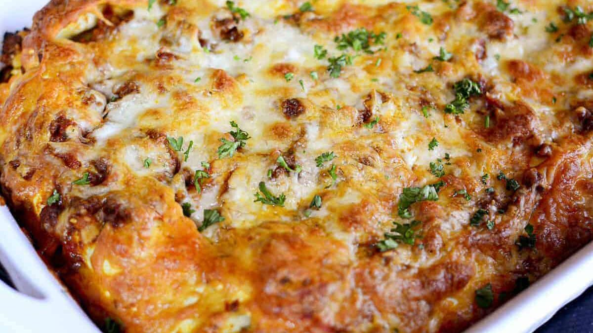 Close-up of a baked lasagna in a white dish. The top is golden brown with melted cheese and small pieces of parsley scattered over it, giving a slightly crispy texture. The dish appears hot, with visible cheese bubbling on the surface.