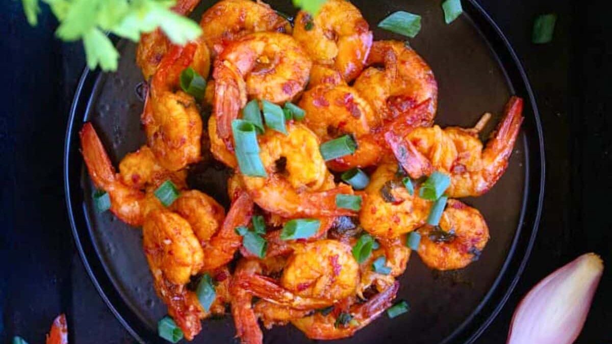 A black plate filled with spicy grilled shrimps garnished with chopped green onions. Fresh green herbs are in the upper left corner and a partial view of a whole red onion is in the lower right corner. The background is dark, making the food vibrant.