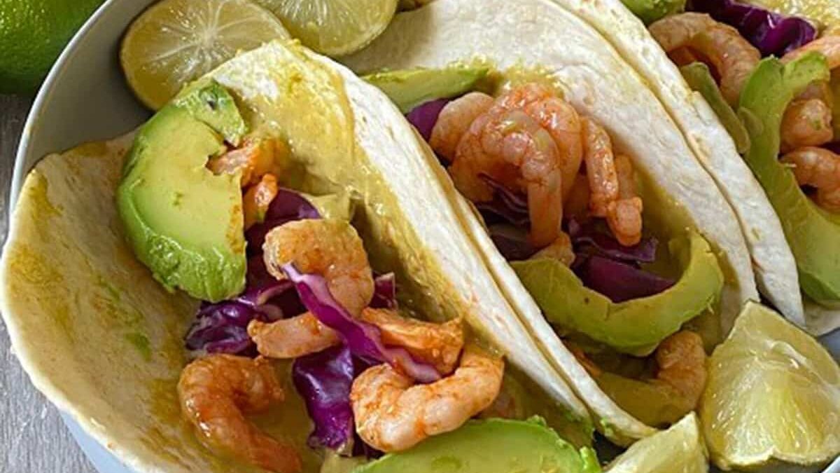 Close-up of shrimp tacos with avocado slices, red cabbage, and lime wedges on a plate. The tacos are filled with grilled shrimp and vibrant toppings, surrounded by lime halves for garnish.