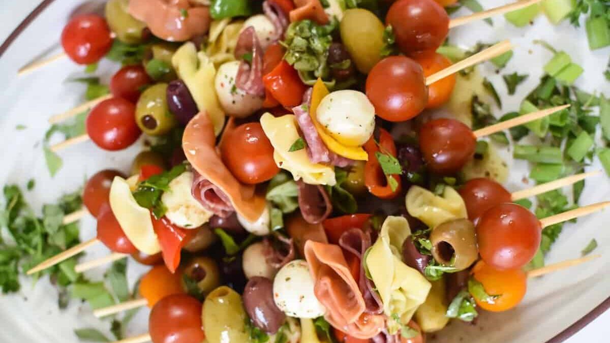 A platter of colorful antipasto skewers featuring cherry tomatoes, mozzarella balls, olives, tortellini pasta, salami slices, and basil. The skewers are arranged on a white dish sprinkled with chopped fresh herbs.