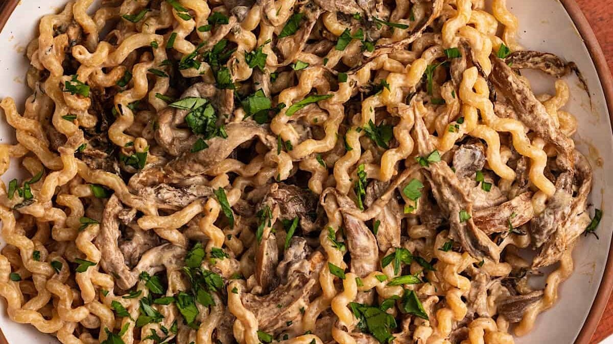 A close-up of a plate of curly pasta mixed with creamy mushroom sauce, topped with chopped parsley. The dish features a rich texture and earthy colors, creating an appetizing appearance.