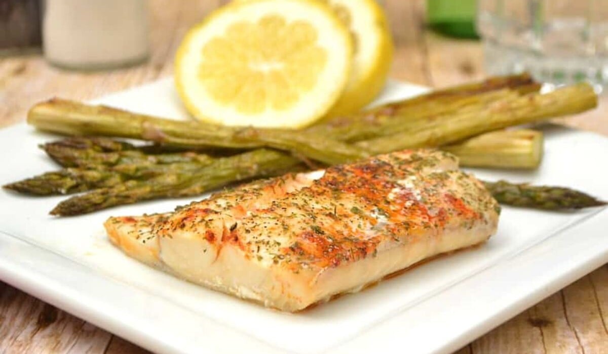 A grilled fish fillet seasoned with herbs sits on a white plate, accompanied by roasted asparagus spears and a slice of lemon in the background.