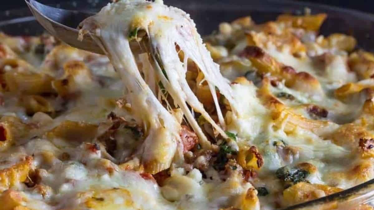 A close-up of a baked pasta dish in a glass casserole dish. The pasta is coated with melted cheese, showing strings of cheese being stretched by a serving spoon, and is mixed with visible herbs and vegetables.