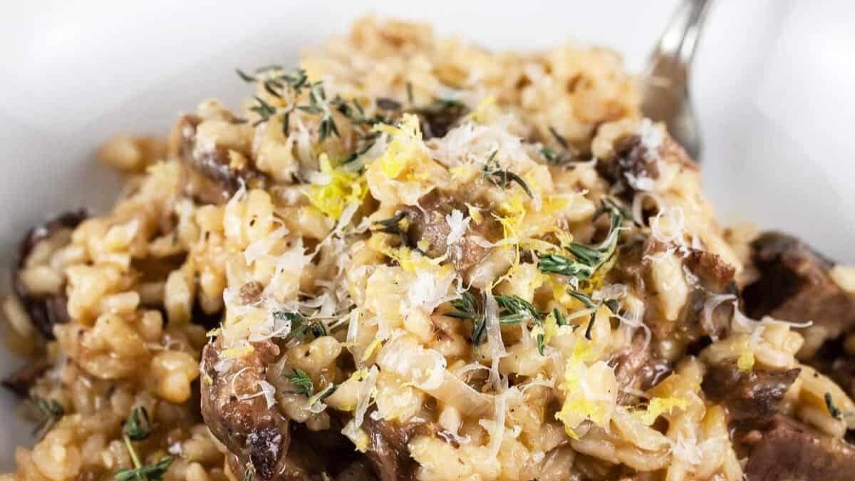 A close-up of creamy mushroom risotto garnished with grated cheese, lemon zest, and fresh thyme on a white plate. A fork is partially visible in the background.
