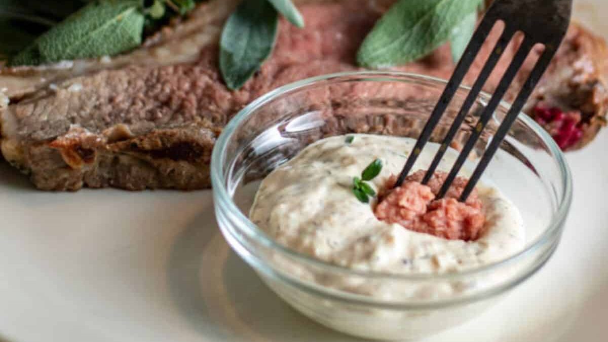 A close-up of a fork dipping into a small bowl of creamy sauce with herbs. In the background, a slice of beef sits on a plate, garnished with fresh green herbs. The setting conveys an appetizing dining experience.