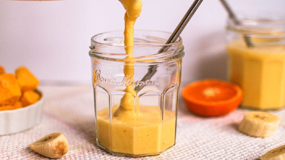 A creamy orange smoothie is being poured into a jar with a metal straw. In the background, there is a halved orange, chunks of mango, and pieces of banana on a fabric surface. Another jar filled with smoothie is visible.