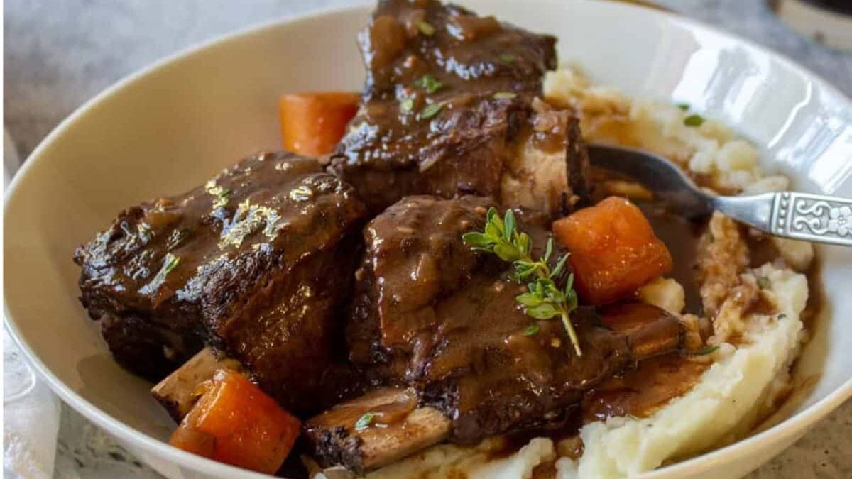 A bowl of braised short ribs in rich gravy served over creamy mashed potatoes, garnished with fresh thyme, and accompanied by chunks of glazed carrots. A spoon rests in the bowl on the side.