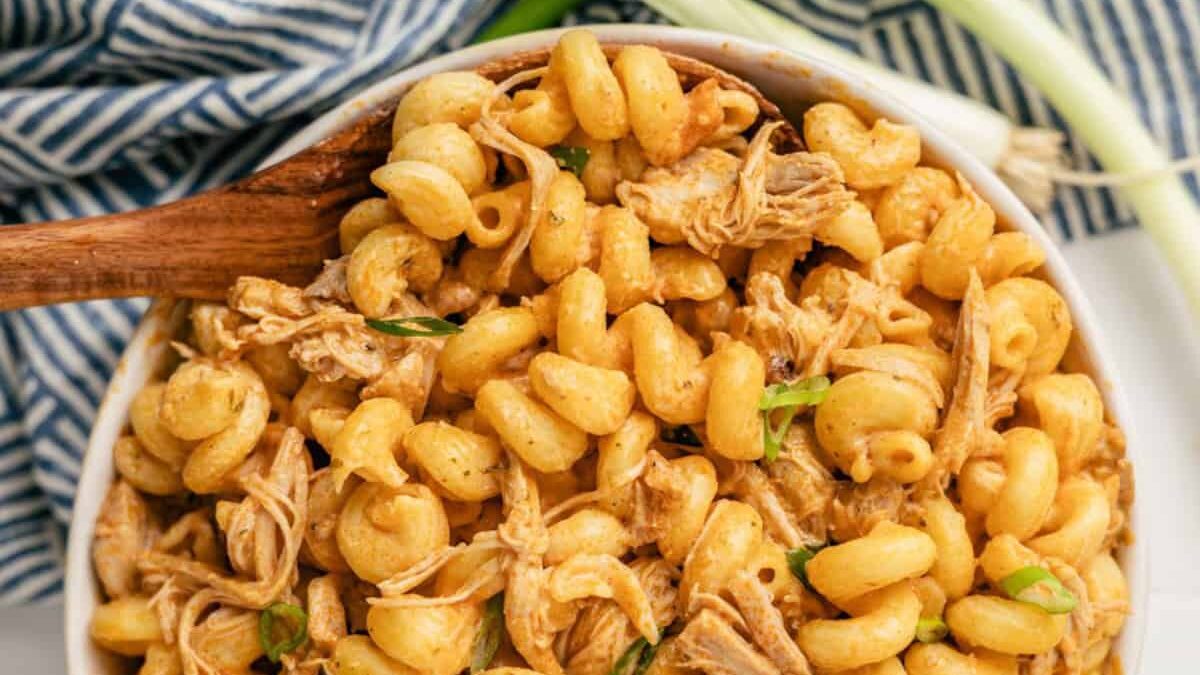 A bowl of creamy pasta with shredded chicken, garnished with chopped herbs. The pasta is a spiral shape, and a wooden spoon is placed to the side. A blue and white striped cloth is partially visible in the background.