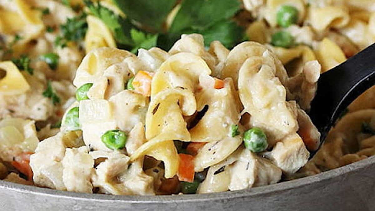 Close-up of a creamy chicken noodle casserole with egg noodles, peas, carrots, and diced chicken, garnished with parsley. A serving spoon is scooping a portion of the dish from the casserole.