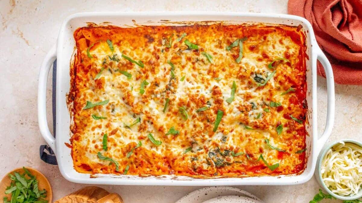 A baked lasagna in a white rectangular dish, topped with melted cheese and garnished with fresh herbs. The dish is surrounded by a small bowl of grated cheese, another bowl of chopped herbs, a woven basket, a plate, and a folded orange cloth.