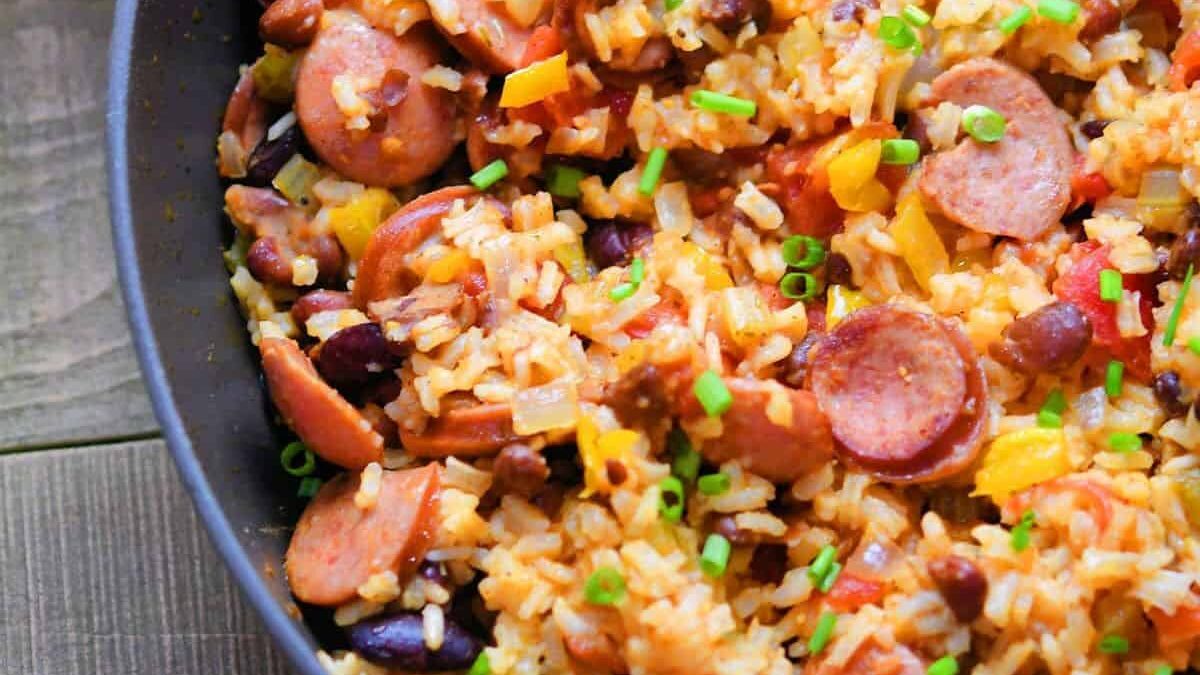 A close-up of a pan filled with rice, sliced sausage, kidney beans, yellow bell peppers, tomatoes, and garnished with chopped green onions. The dish sits on a wooden surface.