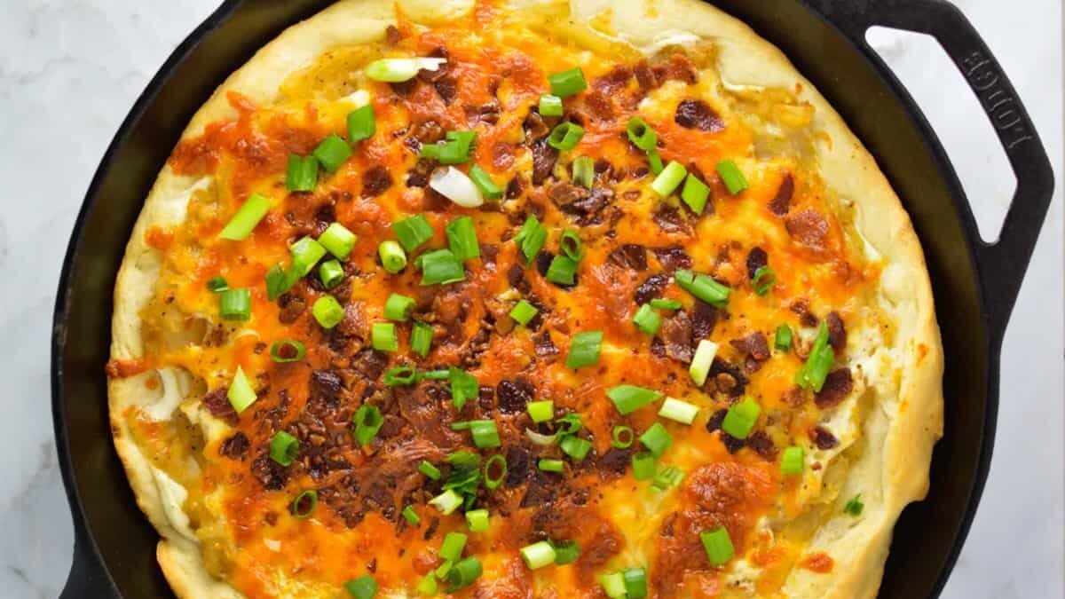 A close-up of a cast-iron skillet containing a baked pizza. The pizza is topped with melted cheese, chopped green onions, and bacon bits. It has a thick, golden-brown crust and is set on a marble countertop.